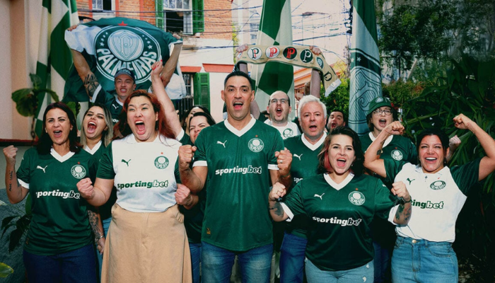 Camisa Palmeiras I 2025 Feminina, cor verde e branca, com gola V retrô branca, estampa especial das taças conquistadas pelo clube e as 12 estrelas dos títulos brasileiros. O escudo oficial do Palmeiras está no lado esquerdo do peito, com os logos da PUMA e Sportingbet, além da frase “De Fato é Campeão” dentro da gola.